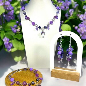 May include: A jewelry set featuring a necklace, bracelet, and earrings with purple beads and mushroom charms. The necklace and bracelet have a silver chain. The earrings are displayed on a white stand with a wooden base. The background has purple flowers.