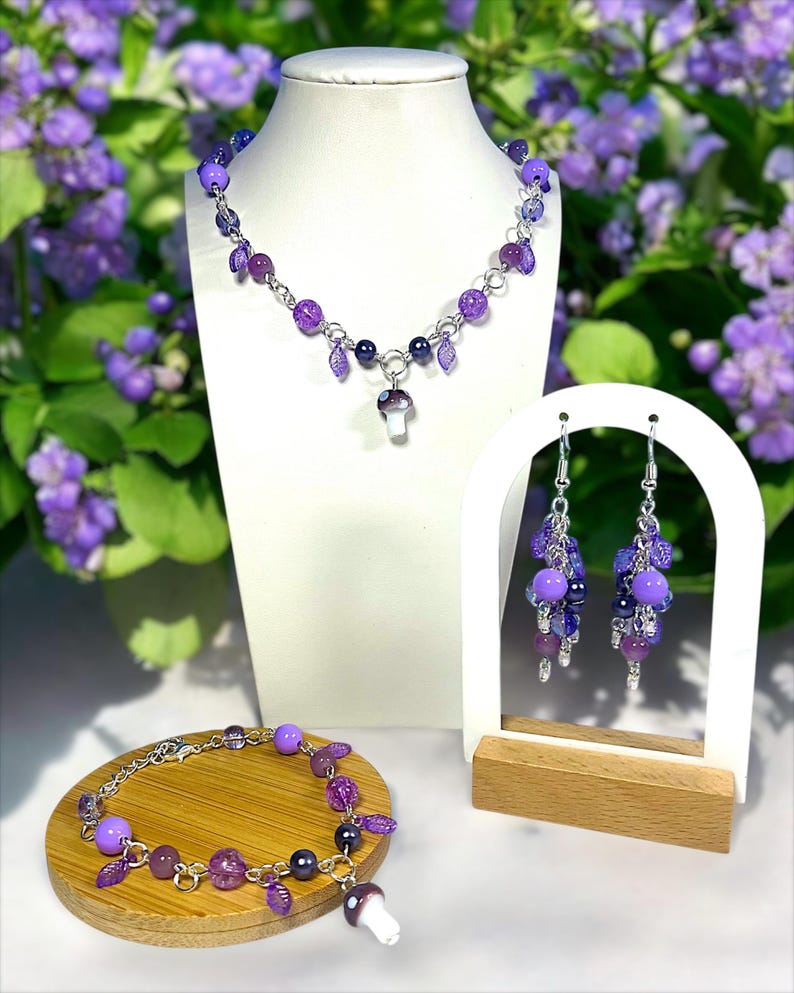 May include: A jewelry set featuring a necklace, bracelet, and earrings with purple beads and mushroom charms. The necklace and bracelet have a silver chain. The earrings are displayed on a white stand with a wooden base. The background has purple flowers.
