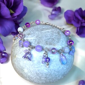 May include: A delicate bracelet with a silver chain and a variety of purple beads and charms. The bracelet features round, iridescent, and teardrop-shaped beads, along with small flower and fairy charms. The background includes purple flowers and a gray stone.