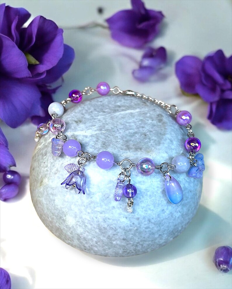 May include: A delicate bracelet with a silver chain and a variety of purple beads and charms. The bracelet features round, iridescent, and teardrop-shaped beads, along with small flower and fairy charms. The background includes purple flowers and a gray stone.