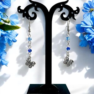 May include: Silver-toned butterfly dangle earrings with blue beads. The earrings are displayed on a black stand against a white background, with blue flowers in the background. The butterfly pendants have detailed wing patterns.