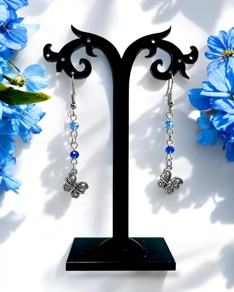 May include: Silver-toned butterfly dangle earrings with blue beads. The earrings are displayed on a black stand against a white background, with blue flowers in the background. The butterfly pendants have detailed wing patterns.