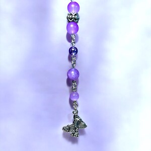 May include: A beaded charm featuring a series of purple beads in varying sizes, connected by silver-colored chain. A silver butterfly charm with rose details dangles at the bottom. The background is a soft, gradient purple.