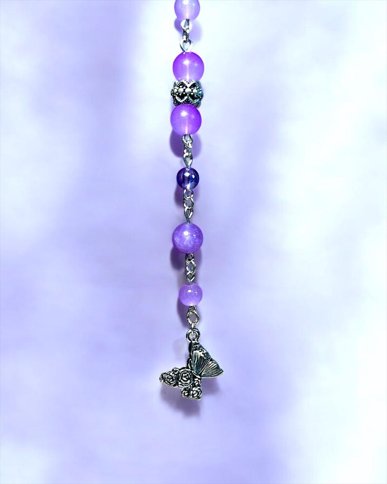 May include: A beaded charm featuring a series of purple beads in varying sizes, connected by silver-colored chain. A silver butterfly charm with rose details dangles at the bottom. The background is a soft, gradient purple.