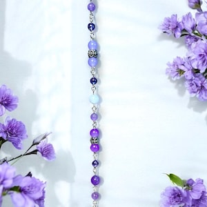 May include: A beaded hair accessory with a gradient of purple beads, ranging from light lavender to deep violet. The beads are strung on a silver chain and culminate in a silver butterfly charm. The accessory is displayed against a white background with purple flowers.