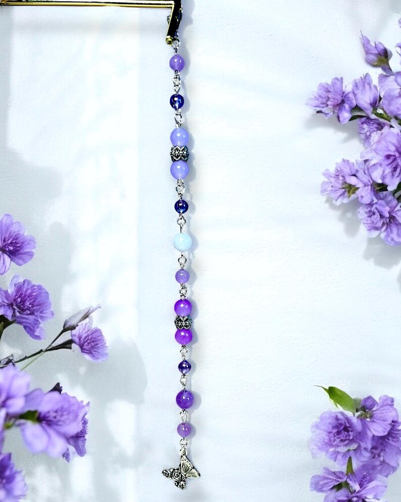 May include: A beaded hair accessory with a gradient of purple beads, ranging from light lavender to deep violet. The beads are strung on a silver chain and culminate in a silver butterfly charm. The accessory is displayed against a white background with purple flowers.