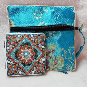 May include: A small, square, white box with a colorful mosaic design. The box is open and has a mirror inside. The box is sitting on top of a blue and gold fabric pouch with a zipper.