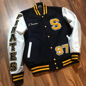 May include: A black and white letterman jacket with yellow accents. The left sleeve has the word "PIRATES" in yellow lettering. The right side features a large yellow "S" and the number "97". The jacket has a ribbed collar, cuffs, and waistband.