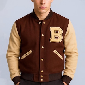 May include: A brown and tan letterman jacket with a large letter "B" patch. The jacket has tan sleeves, a brown body, and gold-colored buttons. The cuffs and collar are brown. Two gold stripes are on the front pockets.