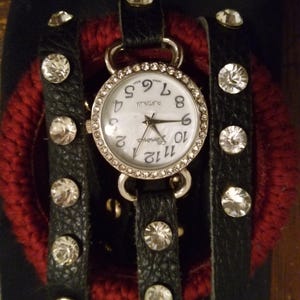 Puede incluir: Un reloj de pulsera Geneva Platinum con esfera blanca y bisel plateado. El reloj tiene una correa de cuero negro con múltiples filas de pedrería redonda y transparente. El reloj está sobre un fondo oscuro con un elemento rojo texturizado.