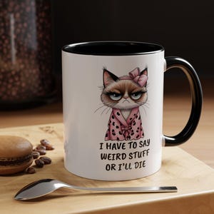 May include: White ceramic mug with a black interior and handle. The mug features a cartoon illustration of a grumpy cat wearing a pink robe and bow. Text on the mug reads: "I HAVE TO SAY WEIRD STUFF OR I'LL DIE."