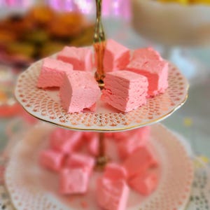May include: A two-tiered white cake stand with a gold trim, holding pink marshmallow squares. The stand is on a white lace doily.