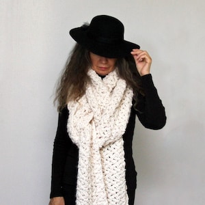 Op de afbeelding: Een witte gehaakte sjaal met lange franjes. De sjaal wordt gedragen door een persoon die een zwarte hoed en zwarte kleding draagt. De tekst "Crochet Pattern" staat onderaan de afbeelding.
