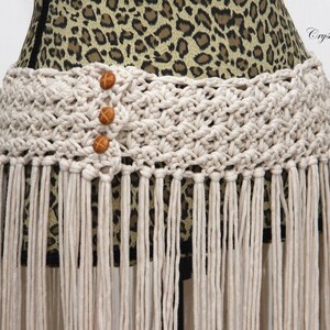 May include: A white crocheted belt with fringe and three brown wooden buttons. The belt is on a leopard print fabric background. Crystal Bear Designs PATTERN.