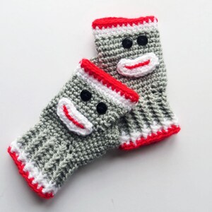 May include: A pair of gray crocheted fingerless gloves with a monkey face design. The gloves have red and white trim and black button eyes.