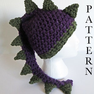 May include: Purple and green crocheted dragon hat with a long tail. The hat has a pointed top and the tail is curled up at the end. The hat is on a white mannequin head. The text "PATTERN" is visible in the background.