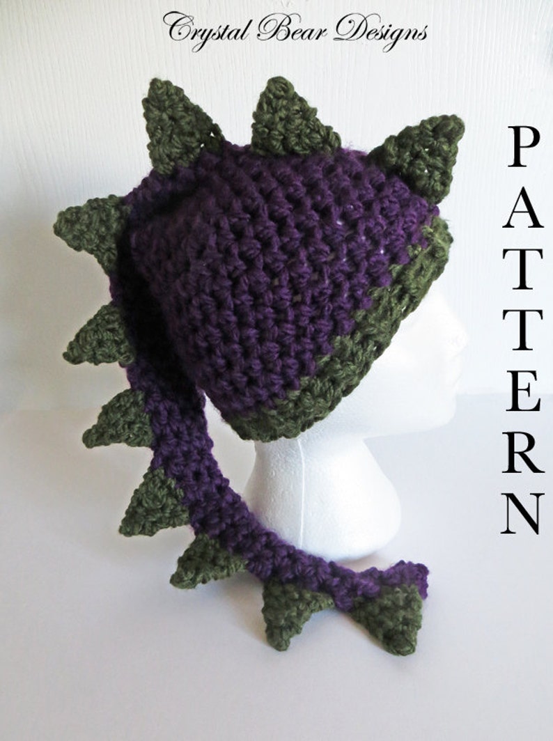 May include: Purple and green crocheted dragon hat with a long tail. The hat has a pointed top and the tail is curled up at the end. The hat is on a white mannequin head. The text "PATTERN" is visible in the background.