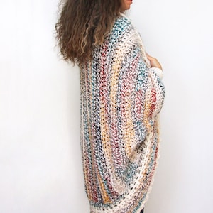 May include: A multi-colored crochet cardigan with a long, flowing design. The cardigan is made with a variety of colors, including white, beige, brown, red, and blue. The cardigan is shown being worn by a person with long, curly hair.