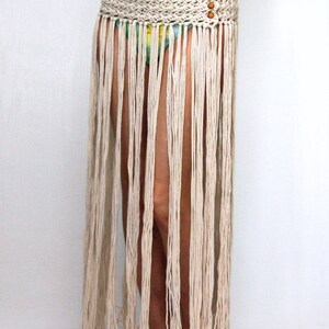 May include: A white crocheted skirt with a fringe hem. The skirt is worn over a floral print swimsuit. The skirt has a woven belt with wooden beads.