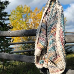 May include: A multi-colored crocheted throw blanket with stripes of teal, red, yellow, and brown hanging on a wooden fence post. The blanket is draped over the post and has a white border.