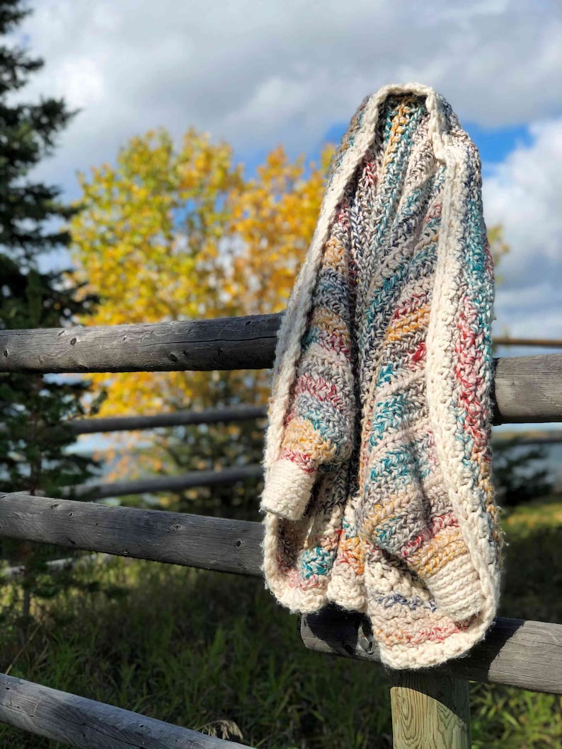 May include: A multi-colored crocheted throw blanket with stripes of teal, red, yellow, and brown hanging on a wooden fence post. The blanket is draped over the post and has a white border.