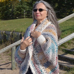 May include: A woman wearing a multi-colored crochet cardigan with a white background. The cardigan has a long, open front and long sleeves. The woman is wearing blue jeans and has long, gray hair.