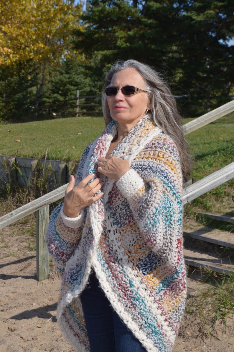 May include: A woman wearing a multi-colored crochet cardigan with a white background. The cardigan has a long, open front and long sleeves. The woman is wearing blue jeans and has long, gray hair.
