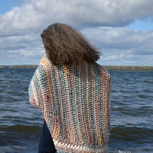 May include: A person wearing a colorful striped crochet cardigan with a white border, standing on a beach with waves crashing behind them.