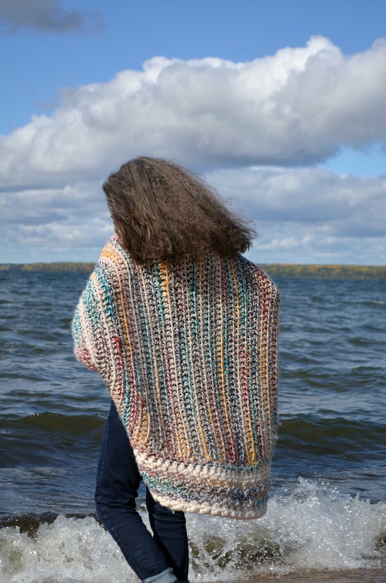 May include: A person wearing a colorful striped crochet cardigan with a white border, standing on a beach with waves crashing behind them.
