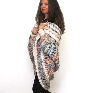 May include: A woman wearing a multi-colored crochet cardigan with a long, flowing design. The cardigan features a variety of colors, including white, brown, teal, and red. The cardigan is draped over her shoulders and arms, and she is wearing black leggings and boots. The text "Crystal Bear Designs PATTERN" is visible at the top of the image.