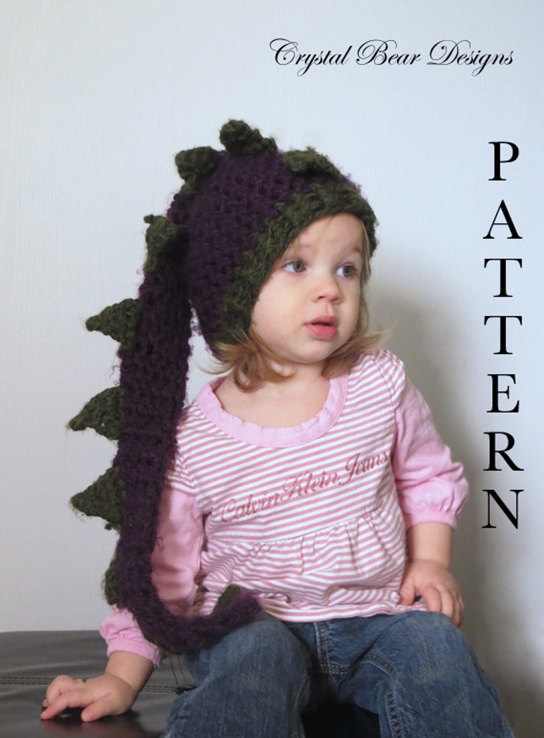 May include: A child wearing a purple and green crocheted dragon hat with a long tail. The hat has a pointed top and a long, flowing tail. The child is wearing a pink and white striped shirt and blue jeans. The text "PATTERN" is written vertically on the right side of the image.
