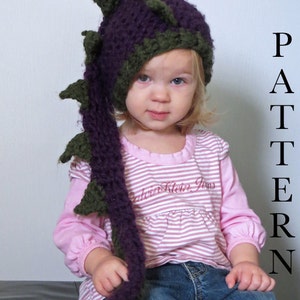 May include: A child wearing a purple and green crocheted dragon hat with a long tail. The hat has a pointed top and spikes. The child is wearing a pink and white striped shirt and blue jeans.