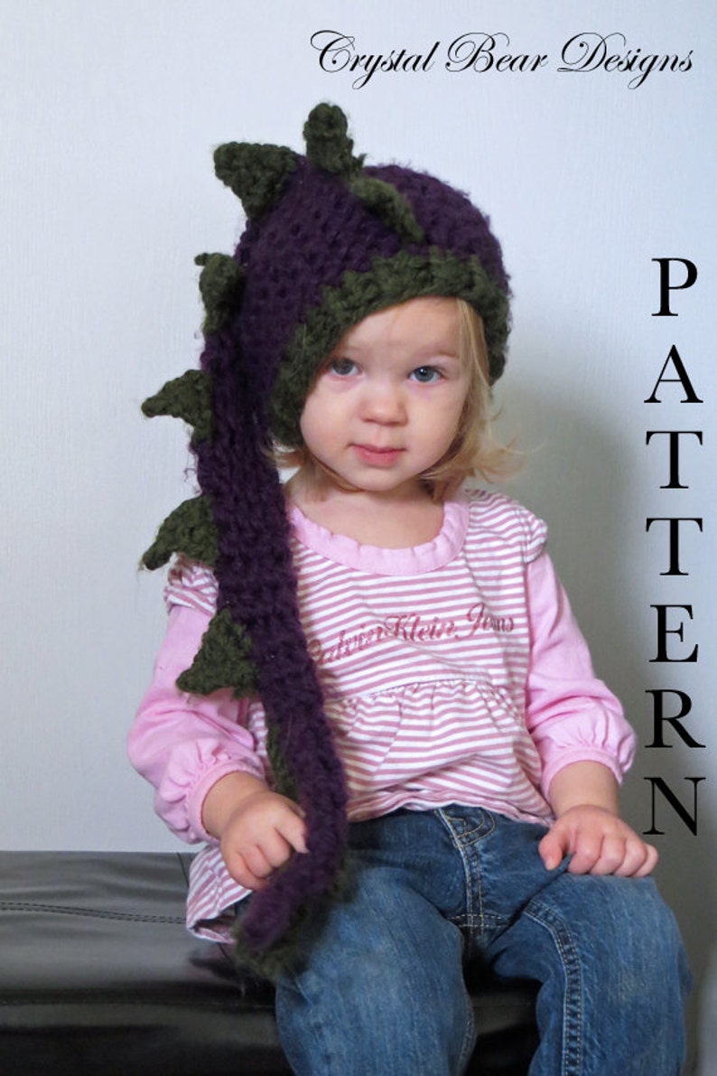 May include: A child wearing a purple and green crocheted dragon hat with a long tail. The hat has a pointed top and spikes. The child is wearing a pink and white striped shirt and blue jeans.
