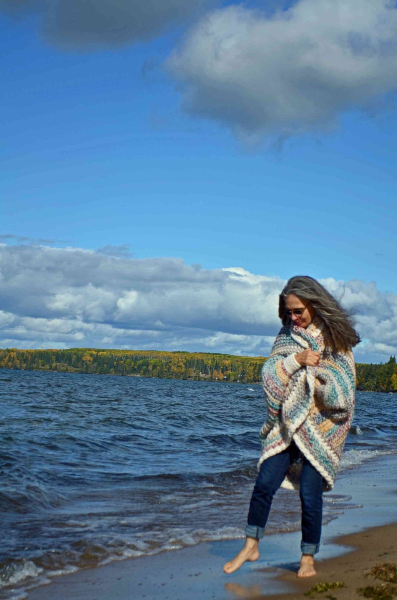 May include: A woman wearing a colorful striped crochet cardigan walks along a sandy beach with blue water and a cloudy sky in the background.