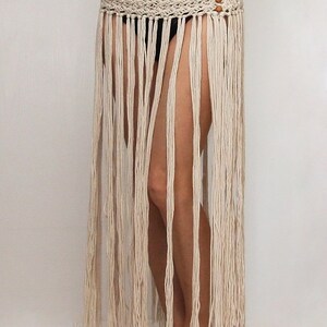 May include: A white crochet skirt with a fringe hem and wooden beads. The skirt is worn by a person with bare legs and feet.