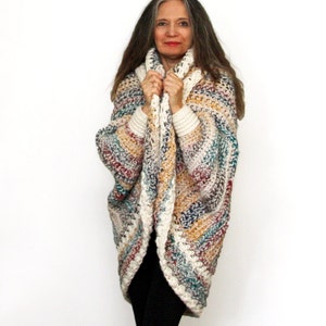 May include: A woman wearing a multi-colored crochet cardigan with a white trim. The cardigan is open in the front and the woman is holding the edges of the cardigan in her hands. The cardigan has a striped pattern with shades of white, brown, blue, and red.