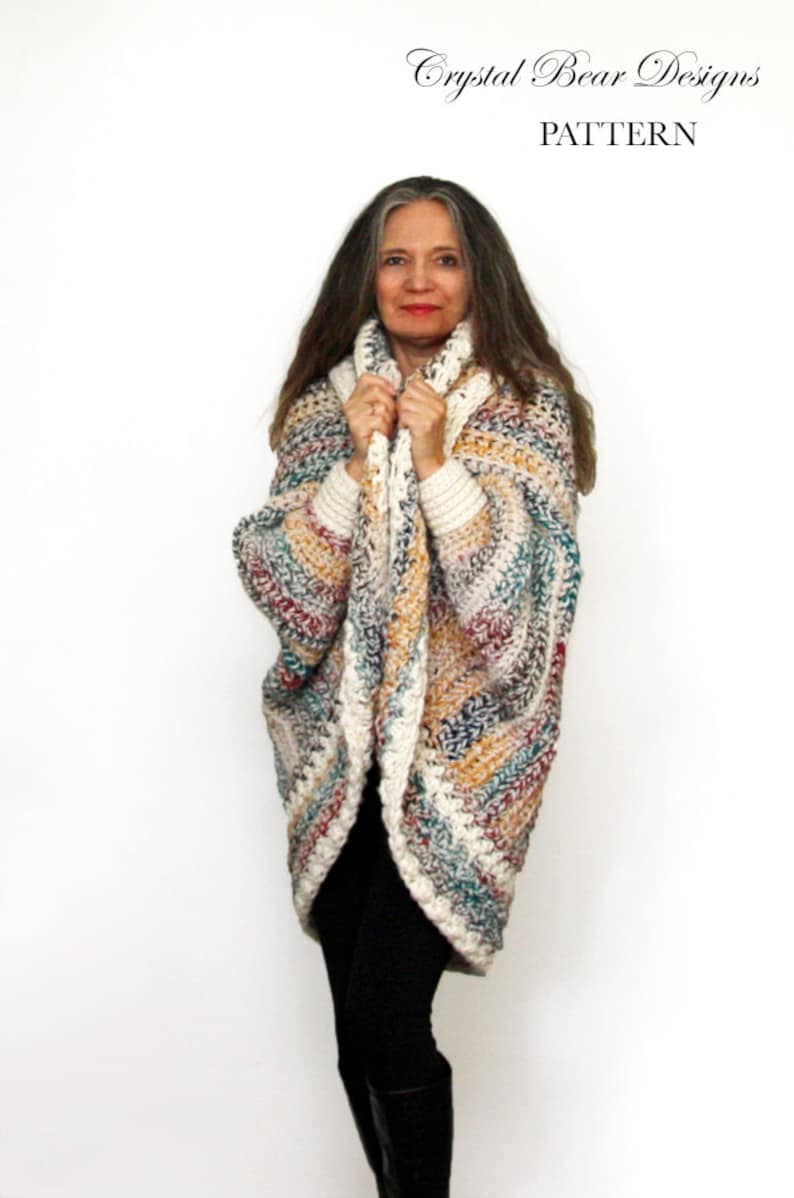 May include: A woman wearing a multi-colored crochet cardigan with a white trim. The cardigan is open in the front and the woman is holding the edges of the cardigan in her hands. The cardigan has a striped pattern with shades of white, brown, blue, and red.
