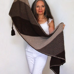 Puede incluir: Un chal de crochet a rayas marrones, beige y blancas con borlas. El chal está drapeado sobre los hombros de una persona.