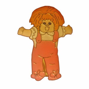 May include: A vintage-style pin featuring a cartoon girl with red hair, wearing a pink dress and overalls. The pin is made of metal and has a gold finish.