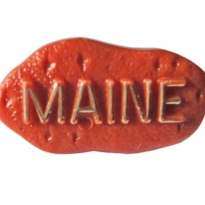 Könnte beinhalten: Ein roter, kartoffelförmiger Souvenir mit dem Wort "MAINE" in goldenen Buchstaben.