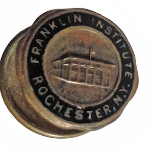 May include: A gold-coloured pin with a black border and the text "Franklin Institute Rochester, N.Y." The pin features a building with a flag on top.