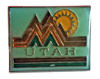 May include: A green enamel pin with a gold outline. The pin features a stylized mountain range with a yellow sun above it. The word "UTAH" is written in gold letters below the mountains.