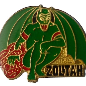 May include: A green and red enamel pin featuring a cartoon devil with wings and the word "ZOLTAH" in black letters.
