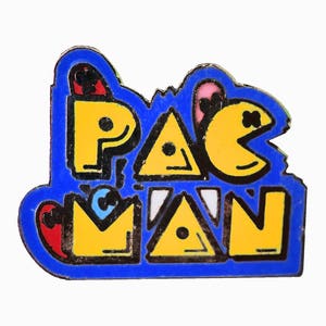 May include: A blue enamel pin with the word "PAC-MAN" in yellow letters with black outlines. The letters are stylized and the "A" is a triangle. The "C" is a Pac-Man character with a pink mouth and black eyes.