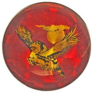 May include: A round, red, glass paperweight with a gold winged horse design. The horse is flying with its wings spread wide. The background is a red, textured surface.