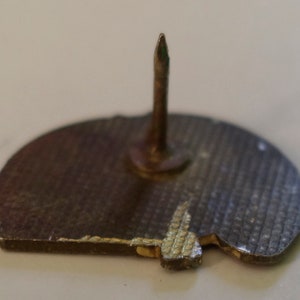 May include: The back of a silver pin with a textured surface and a pointed pin on the back.