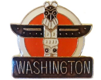 May include: A silver and black enamel pin featuring a stylized totem pole with a red and white circle behind it. The text "WASHINGTON" is on a black banner below the totem pole.