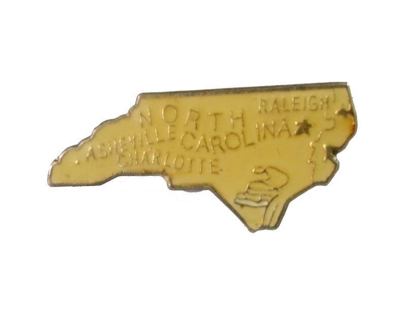 Op de afbeelding: Een gele metalen pin in de vorm van de staat North Carolina. De pin heeft de woorden "North Carolina" en de steden "Raleigh", "Asheville" en "Charlotte" erop geschreven.