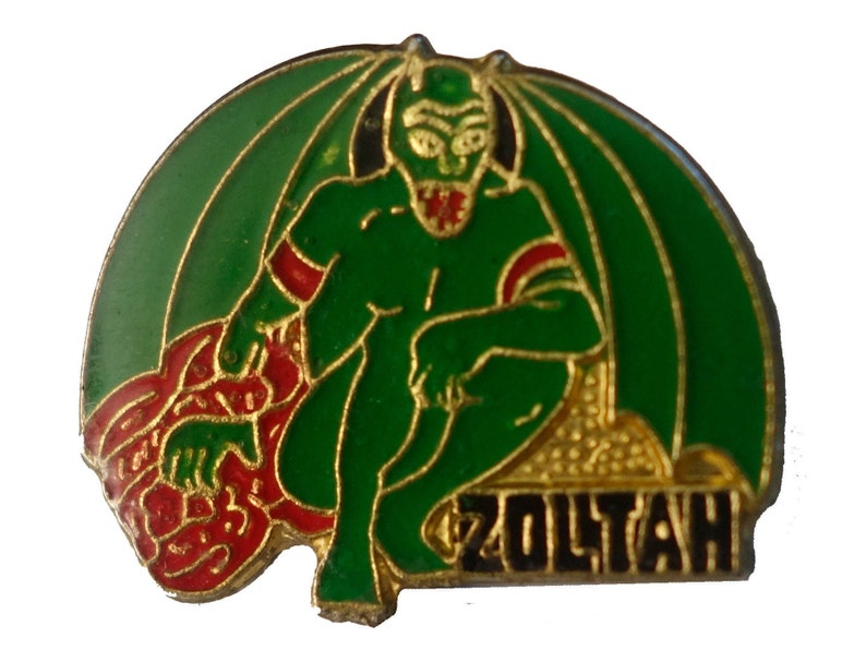 May include: A green and red enamel pin featuring a cartoon devil with wings and the word "ZOLTAH" in black letters.