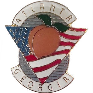 May include: A gold-colored pin with a white background and the words "Atlanta Georgia" in a circle around a peach and an American flag. The peach is orange and has a green leaf. The flag is red, white, and blue.